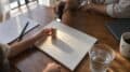 Overhead view of a dining table where an adult hand holds a torch over a white card and wooden block beside a cup of water.